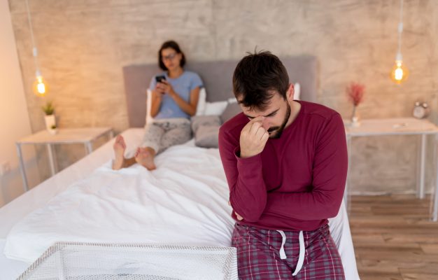 Couple fighting in bedroom, woman using a smartphone in the background while man sitting in the foreground, sad and pensive; relationship and marital problems concept