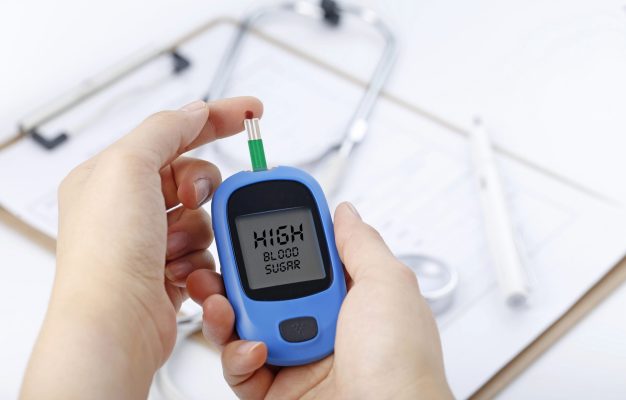 Hand holding a blood glucose meter measuring blood sugar, the background is a stethoscope and chart file